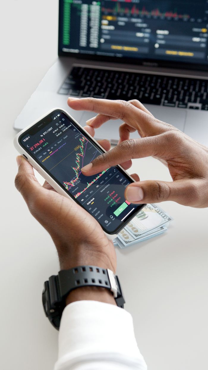 Hands holding a smartphone with a trading app and chart overlays, with dollar bills in the background.